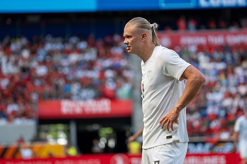 Archive photo: Erling Haaland stands on the pitch wearing a Norway shirt, with a packed stand blurred behind him. Source: Wikimedia Commons